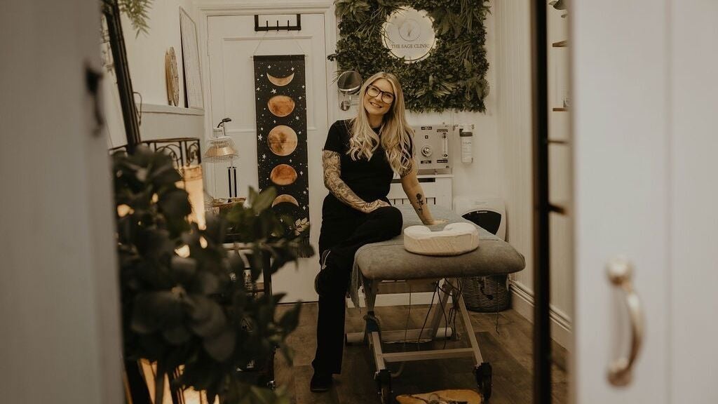 Amiee seated on the treatment couch in her clinic, beside a moon-phase wall hanging and a wall of greenery