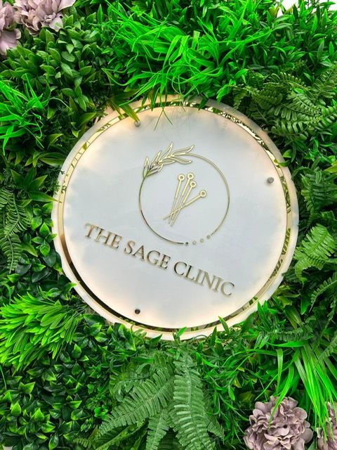 The Sage Clinic gold and ivory sign mounted on a lush greenery wall dotted with pink flowers